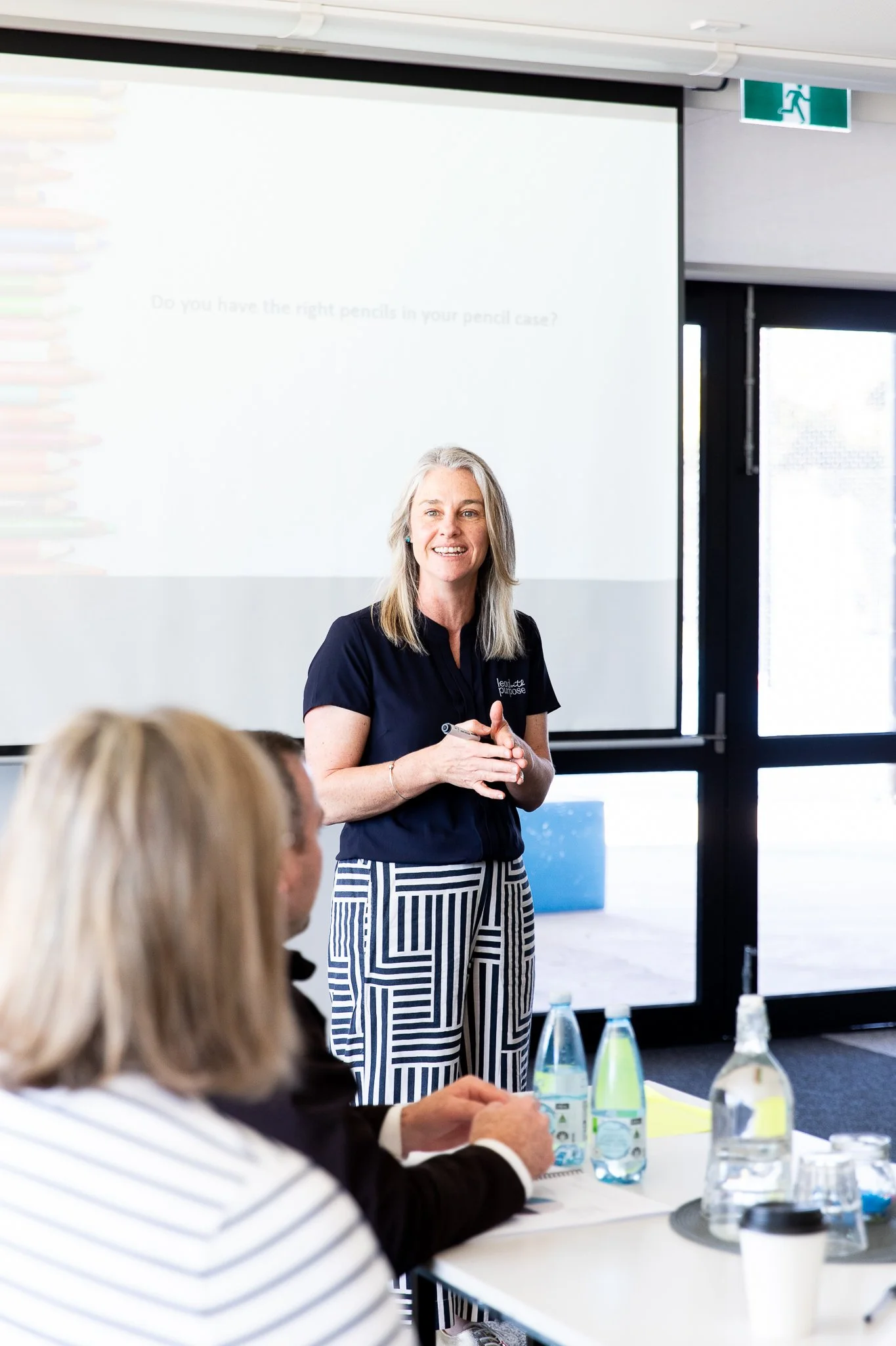 Karissa Cameron, a woman, wearing a navy blue shirt standing in front of a white projection screen speaking to a group in a meeting room.