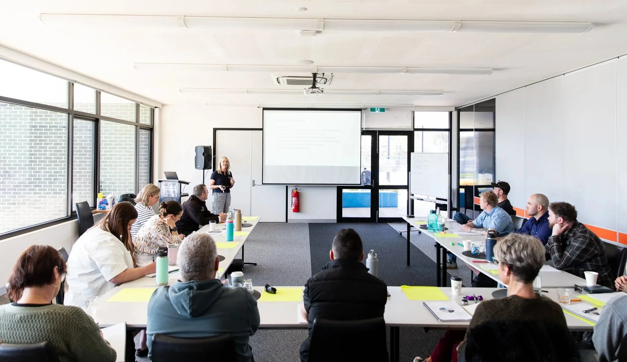 Karissa Cameron, a woman presenter giving a talk to a group of adults seated at tables in a conference room with large windows.