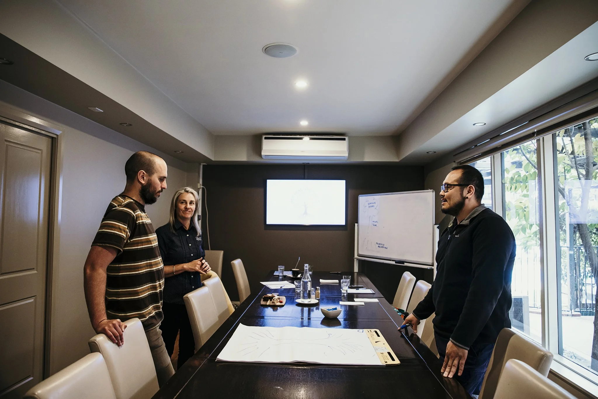 Three people in a conference room preparing for a presentation. Two men and one woman are standing around a long table with documents, water bottles, and glasses. The room has large windows on one side and a whiteboard and TV screen on the back wall.