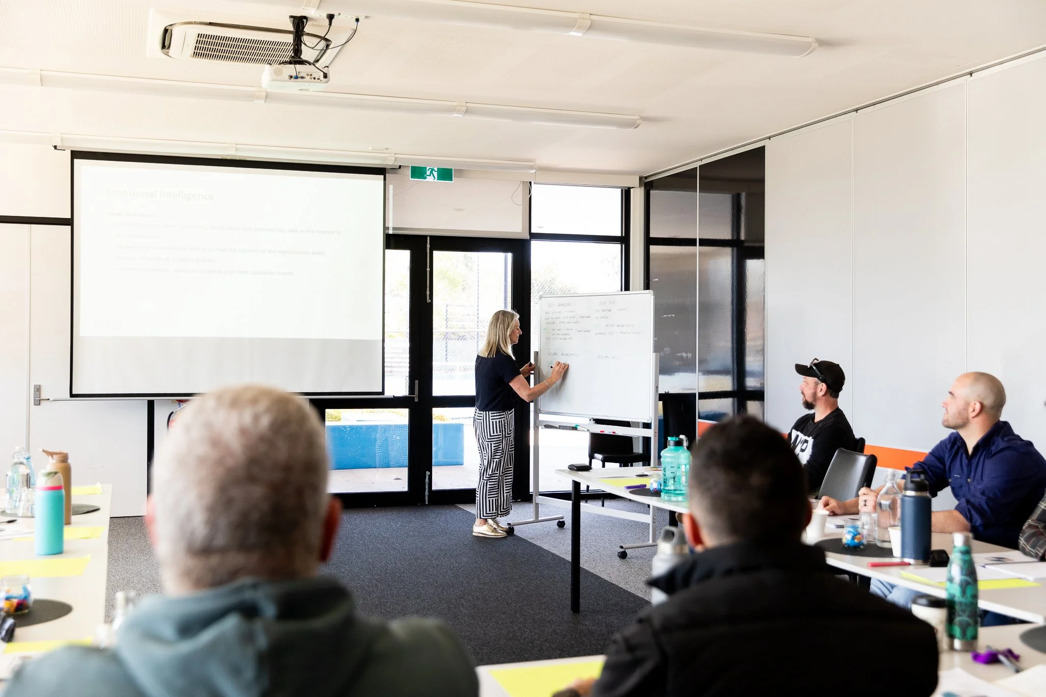 Karissa Cameron, a woman presenting at the front of a classroom or conference room to an audience of four men, with a whiteboard and a projector screen displaying presentation slides.