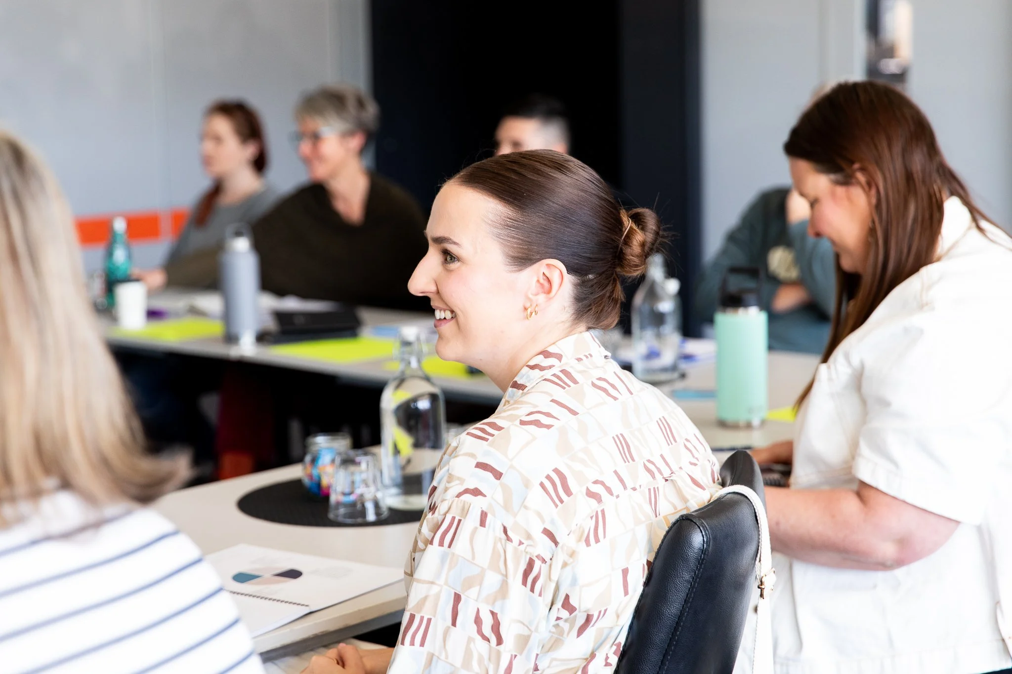 A woman in a leadership workshop with dark brown hair tied in a bun, wearing a patterned shirt, smiling during a meeting in a modern conference room with other women present.
