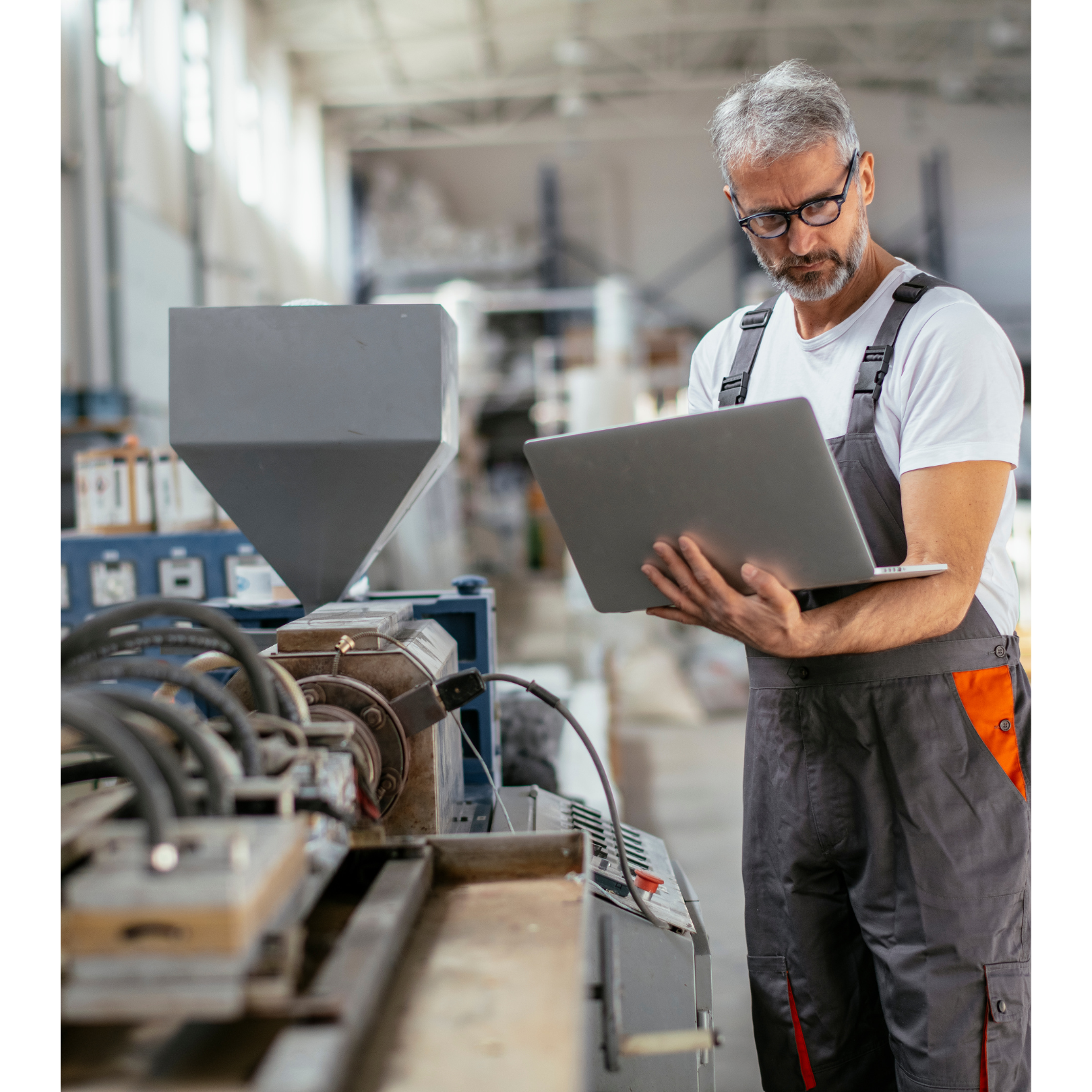 An engineer with gray hair and glasses wearing a white t-shirt and gray overalls standing in a factory or workshop with industrial machinery, holding a laptop and inspecting equipment.