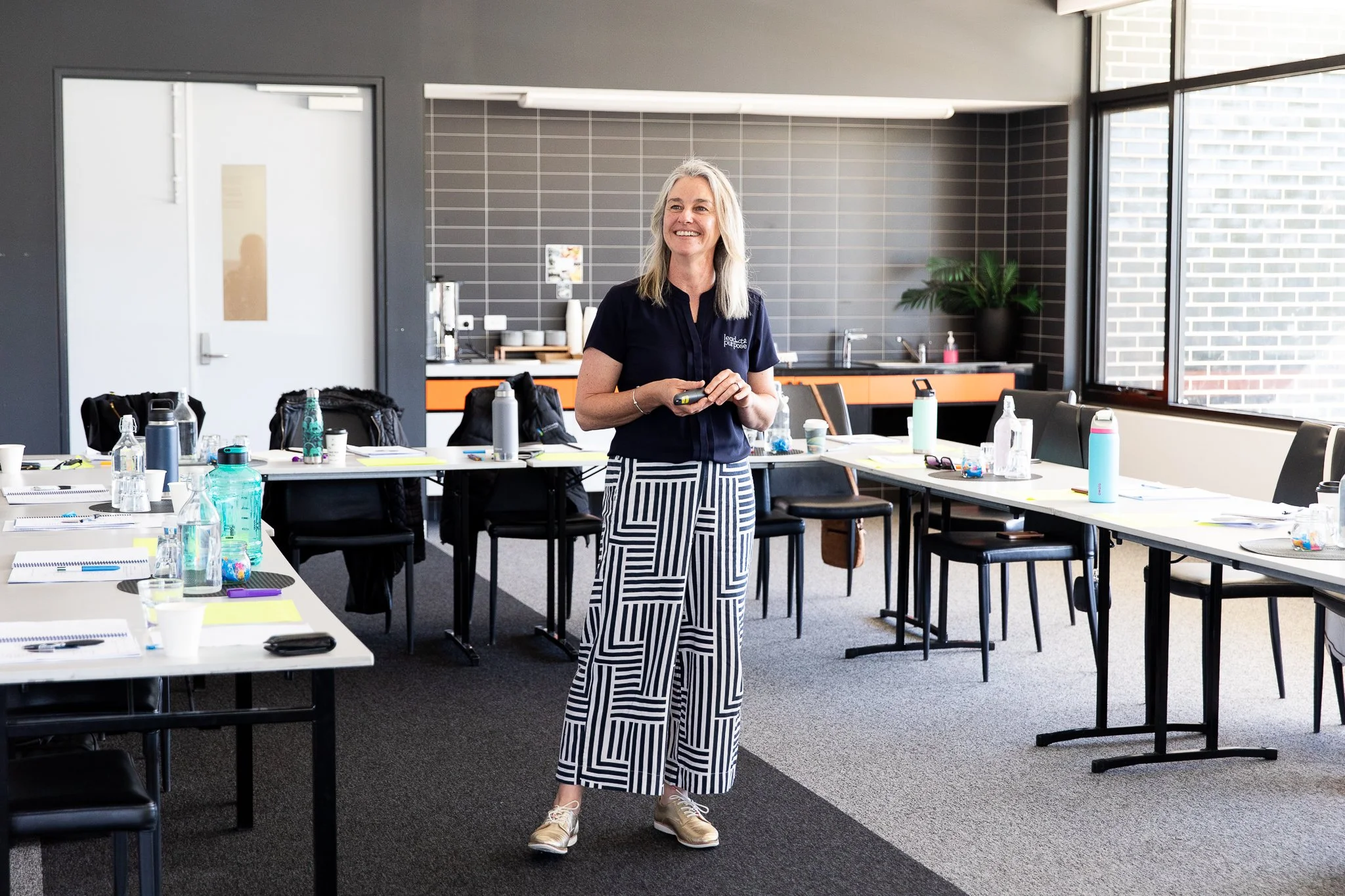 Karissa Cameron, a woman, standing in a conference room, smiling, holding a remote or a small device. The room has tables with water bottles, notebooks, and glasses, and large windows allowing natural light.