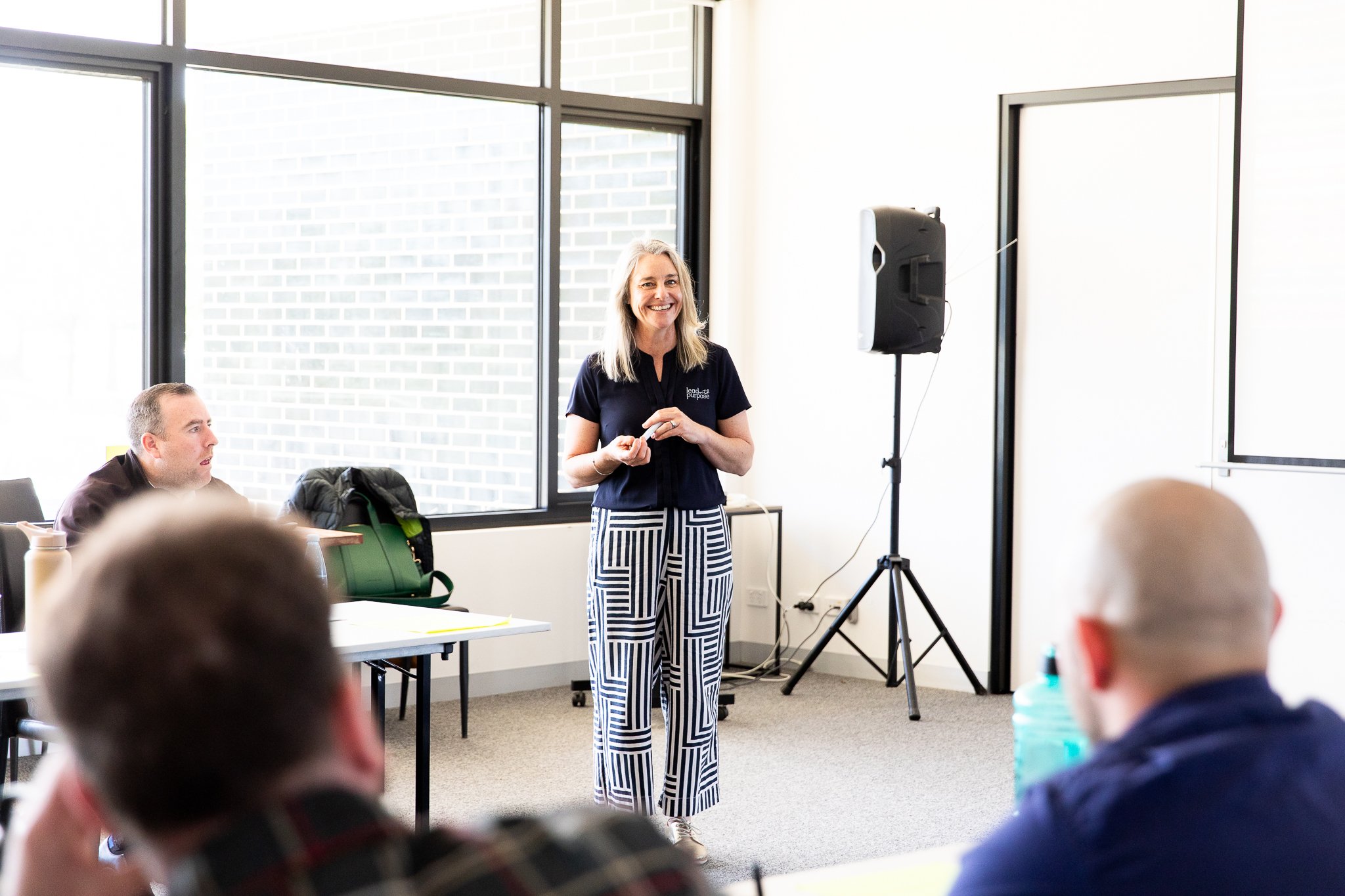 Karissa Cameron giving a presentation to a leadership team in a bright conference room. She is smiling and standing near a speaker, with several seated attendees listening. Large windows are in the background.