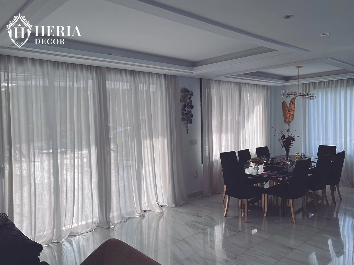 Sala comedor con cortinas blancas, mesa negra con seis sillas negras, lámpara colgante, flores en un jarrón y decoraciones en las paredes.