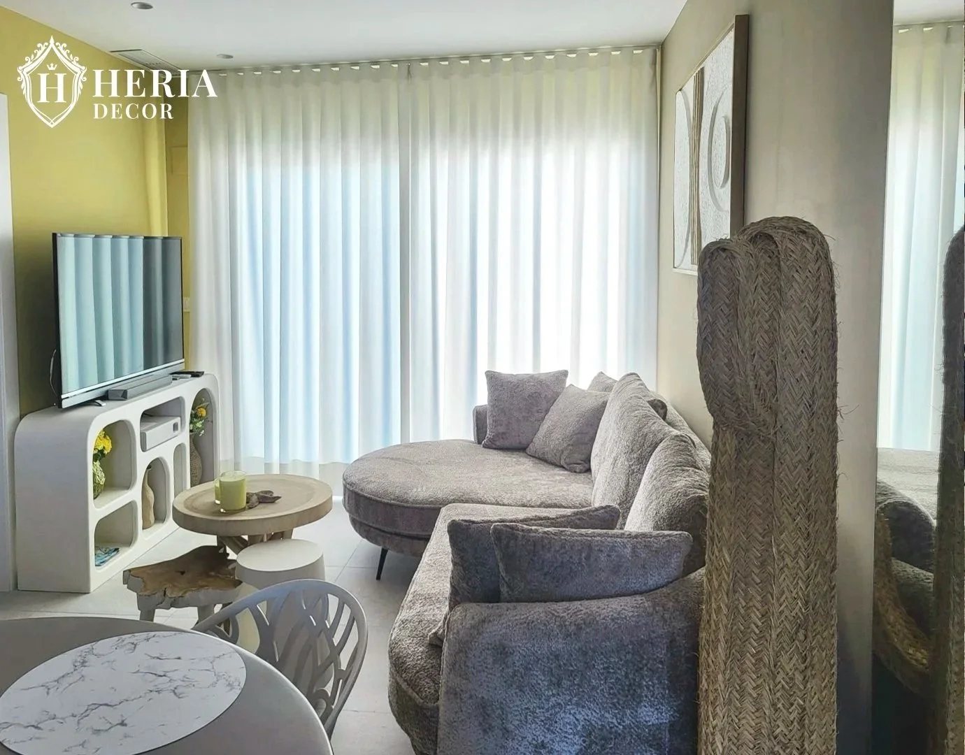 Sala de estar con cortinas blancas, sofá gris con cojines, mesa de centro y televisor en mueble blanco con decoración floral.