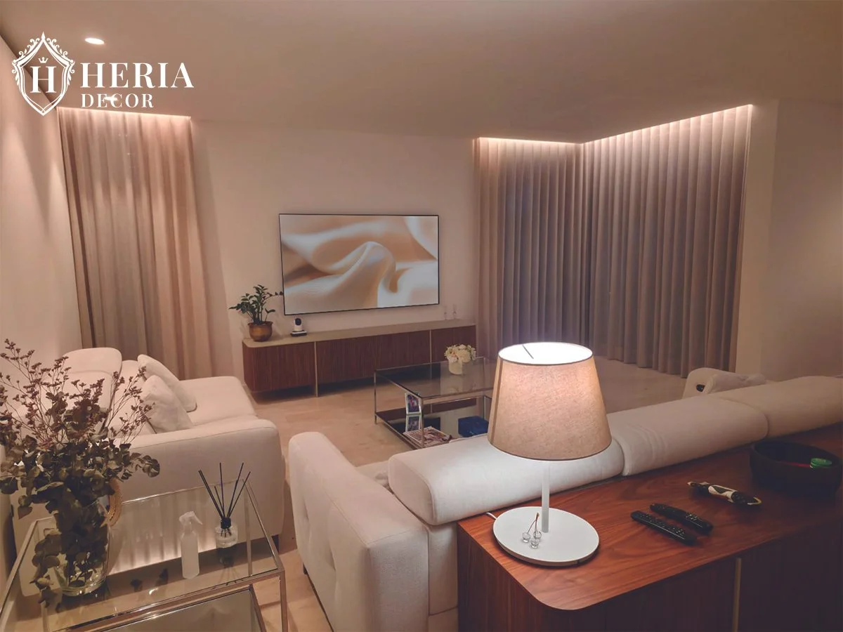 Sala de estar con sillones blancos, una mesa de centro de vidrio, televisión, cortinas largas beige y una lámpara en una mesa de madera.