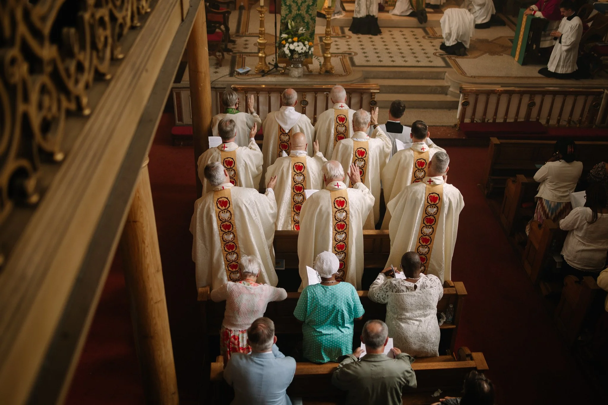 Clergy members wearing white robes with embroidered red hearts and gold decorative patterns, gathered in a mass in a church.