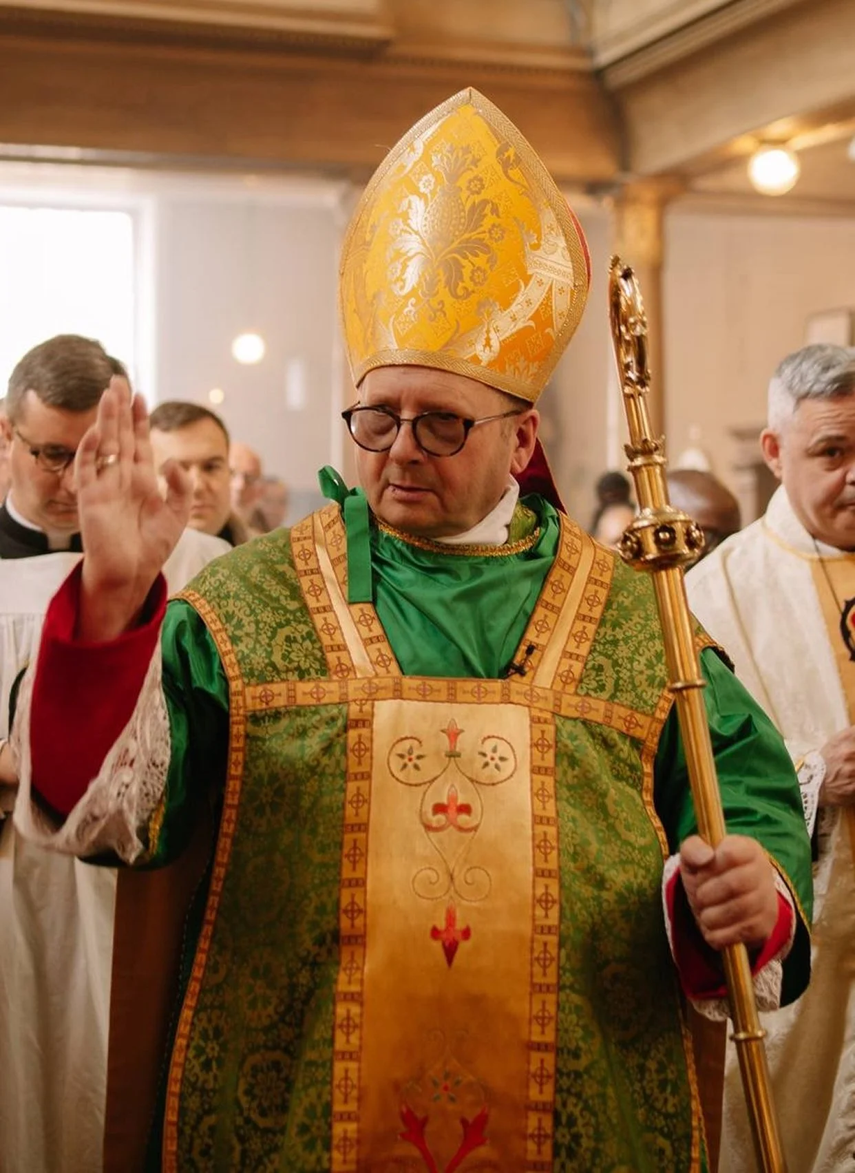 Bishop David in ornate green and gold vestments with a tall, golden mitre on his head, holding a staff, and raising his right hand in blessing during a mass.