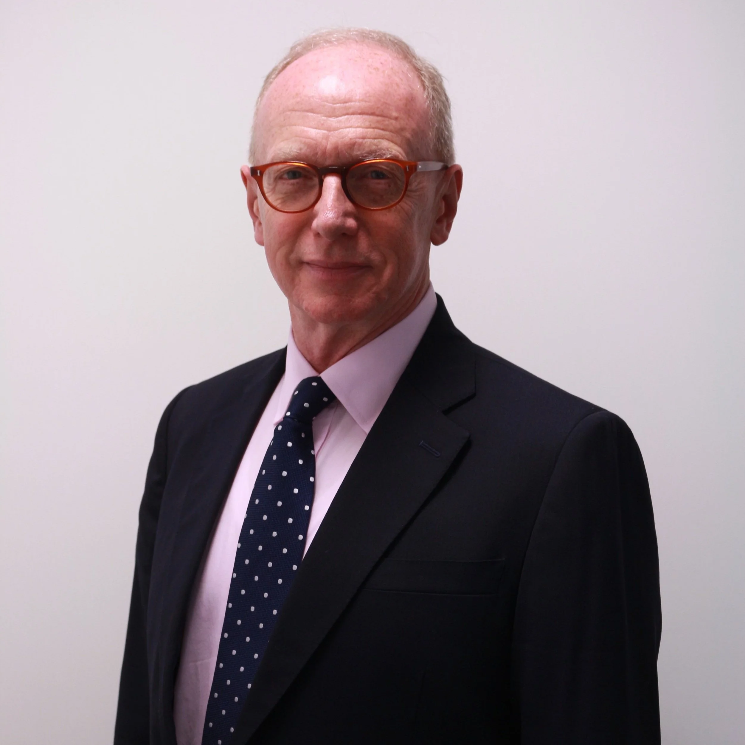 Nicolas professionally dressed with glasses, wearing a black suit, pink shirt, and polka dot tie, standing against a plain white background.