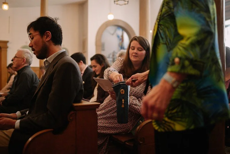 People sitting in pews inside a church, with one woman reaching into a bag to offer her donation and others seated.