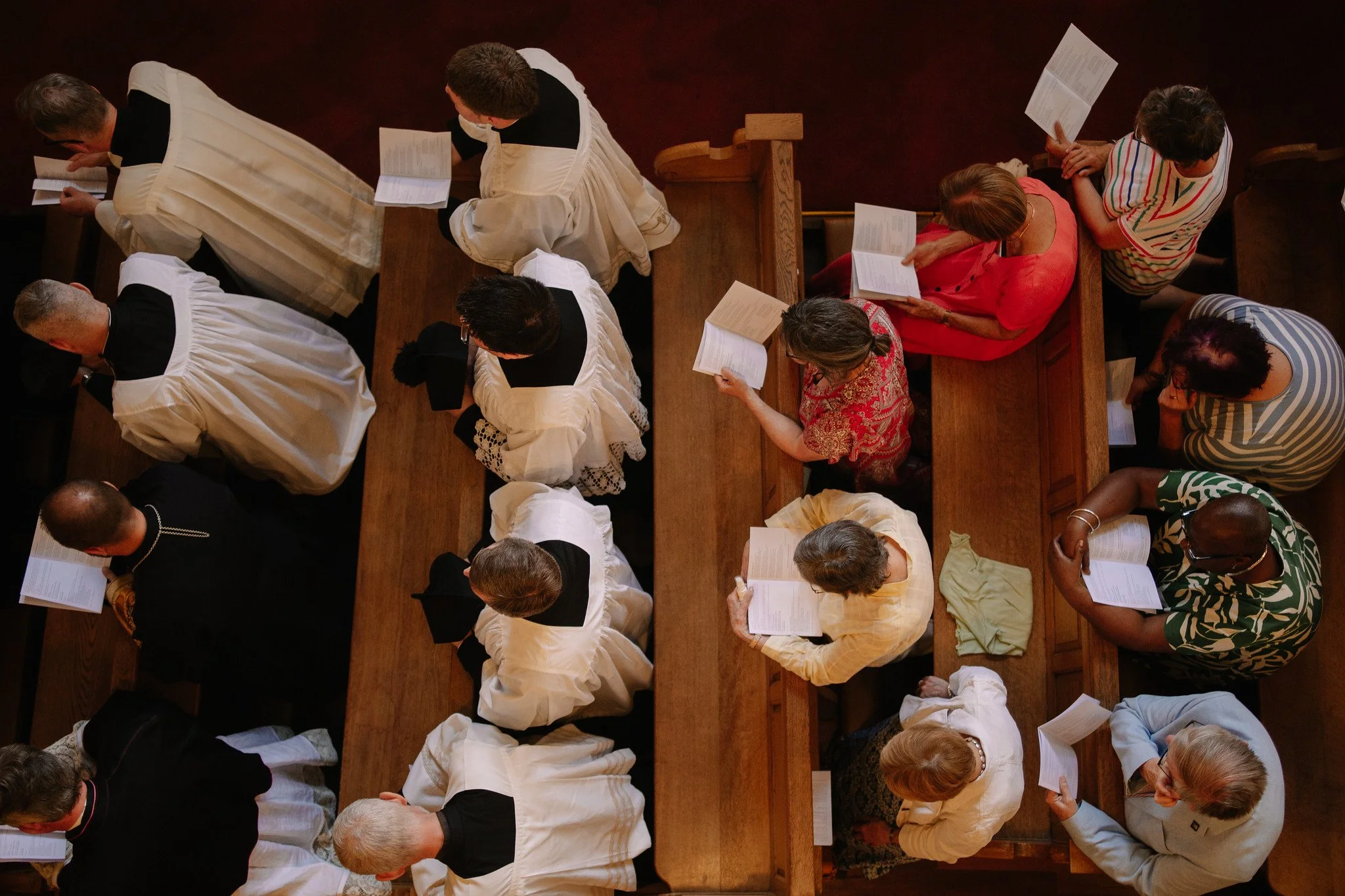 People standing in pews inside a church, reading prayer books, seen from above in a church.