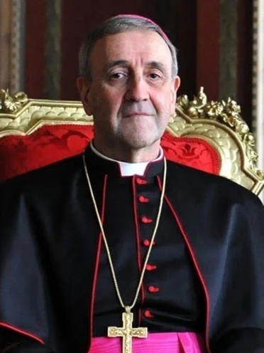 A Catholic bishop sitting on a gold ornate chair, wearing black clerical robes with red accents, a pectoral cross on a gold chain, and a purple sash.