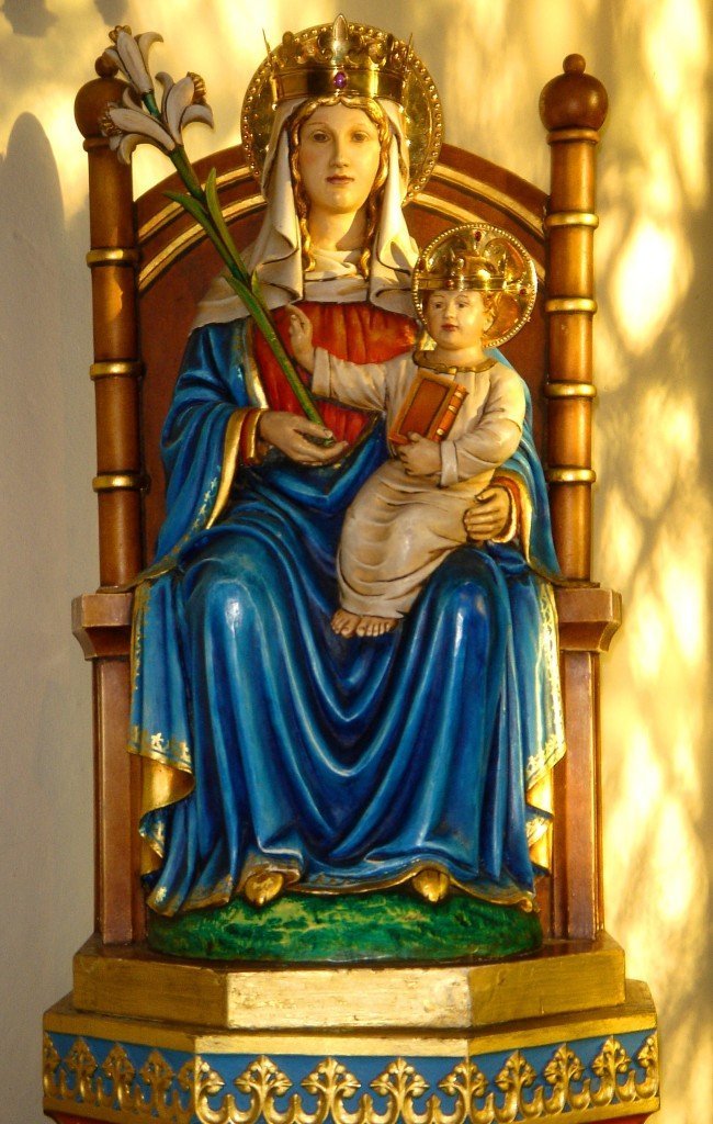Colorful wooden statue of Our Lady and Child Jesus, with Mary wearing a blue robe and a crown, holding lilies and the Child holding a book, both with gold halos, seated on a decorated throne.
