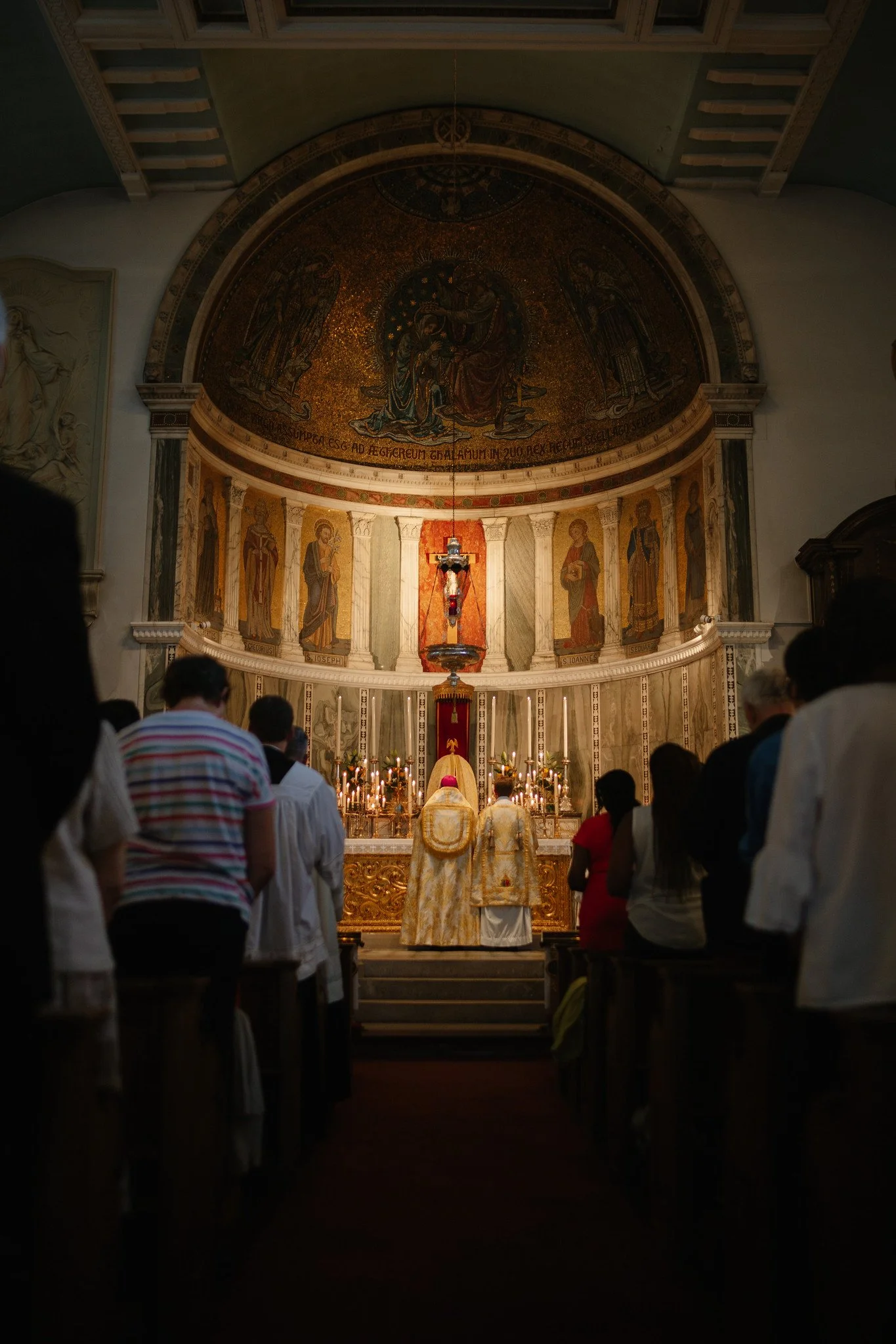 People are gathered at Warwick Street Parish, standing and participating a mass. The altar is decorated with candles, and there are ornate mosaics on the wall behind.