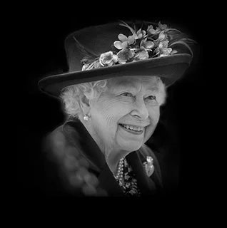 Queen Elizabeth II wearing a large hat decorated with flowers and a black coat with pearls.