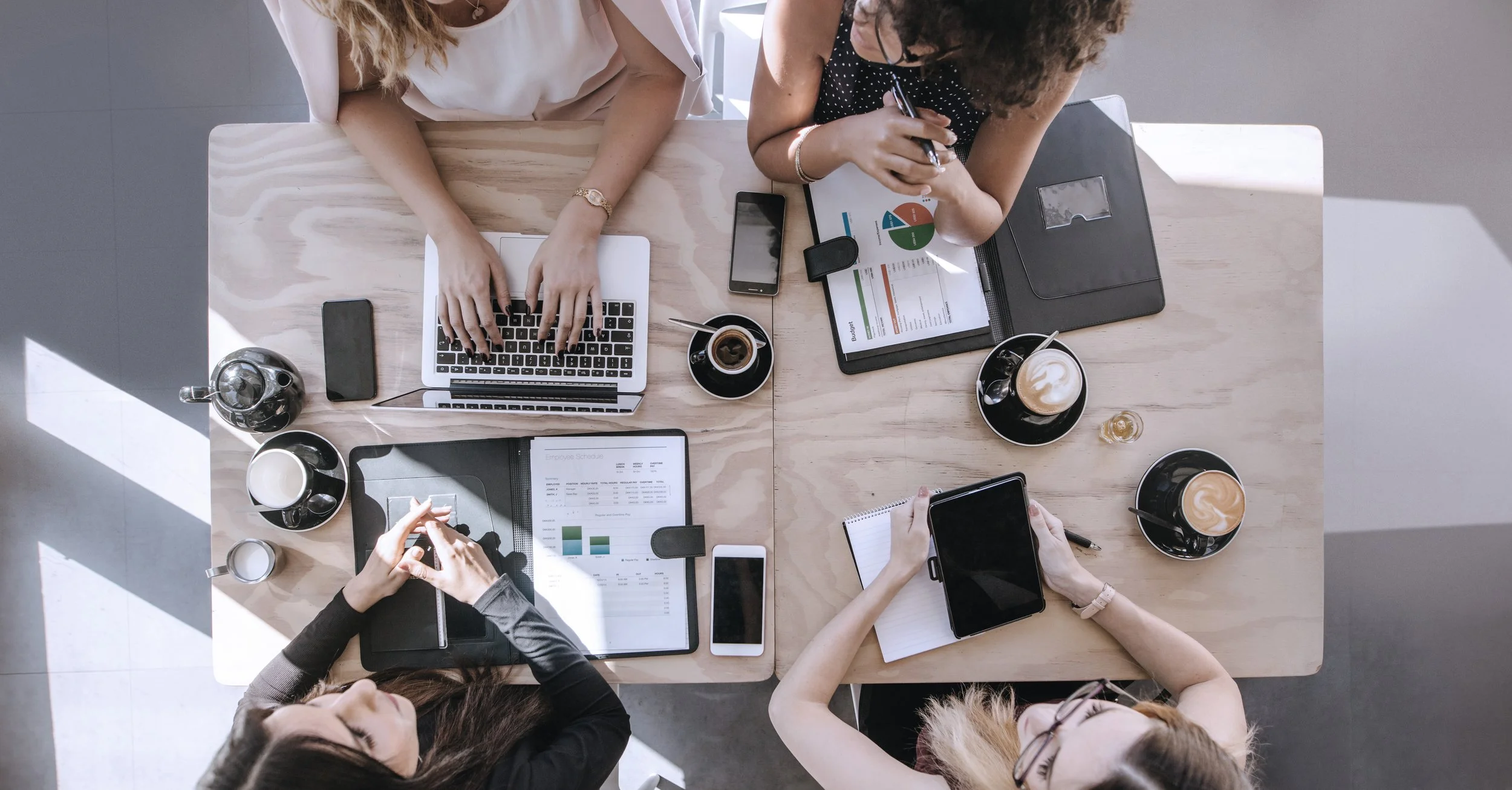 Top-down view of four women seated around a wooden table working with laptops, tablets, notebooks, and smartphones, with coffee cups and notepads on the table.
