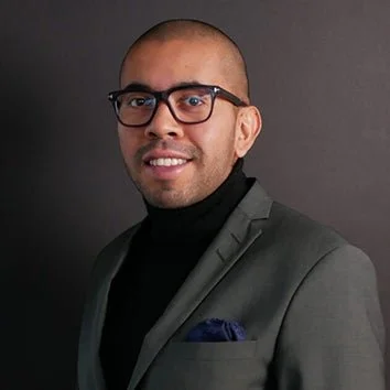 A man with a shaved head wearing black glasses, a gray suit jacket, black turtleneck, and a navy pocket square, posing against a dark background. Ricardo Cuilenheim.