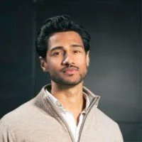 Portrait of a man with dark hair and a beard, wearing a light-colored sweater, standing against a dark background. Sayanthan Kamal.
