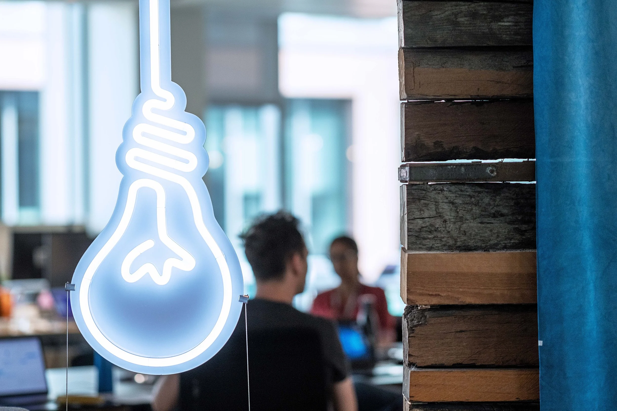 Neon sign of a light bulb and brain outline in a café or workspace, with blurred people in the background and a brick wall on the right.