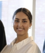 A young woman smiling with her hair tied back, wearing a white shirt, in a professional setting with a window in the background. Nora Emilie Carlsson.