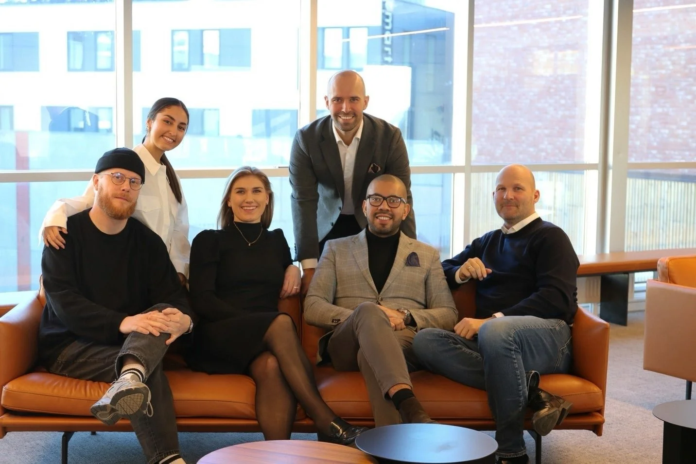 Group of six diverse business professionals in an office lounge, smiling for a photo. Three are seated on a leather sofa, and three stand behind them. Large windows with city buildings in the background.