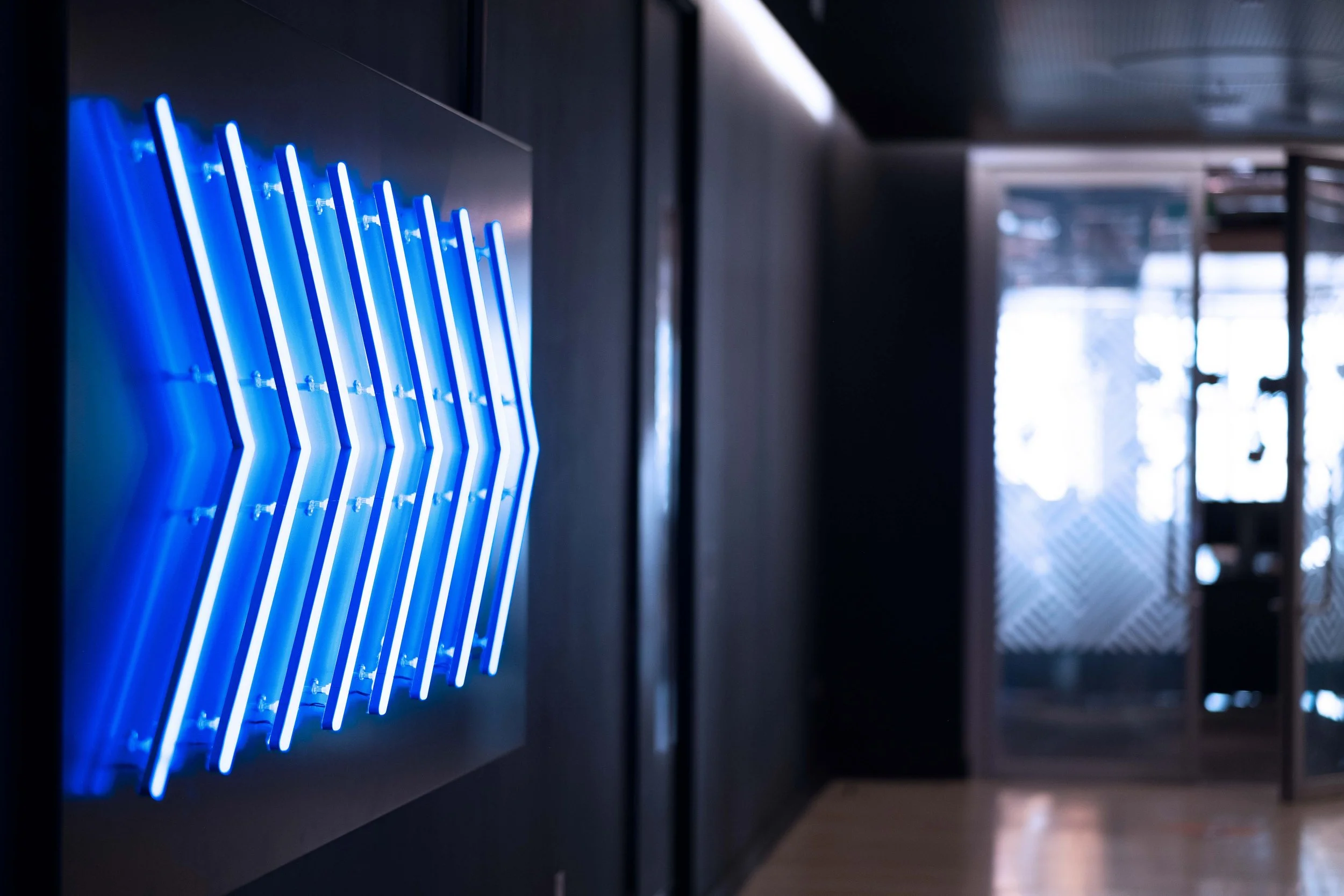 Blue neon artwork with vertical lines in a geometric pattern on a dark wall in an indoor space, with a window or glass door showing outdoor scenery in the background.