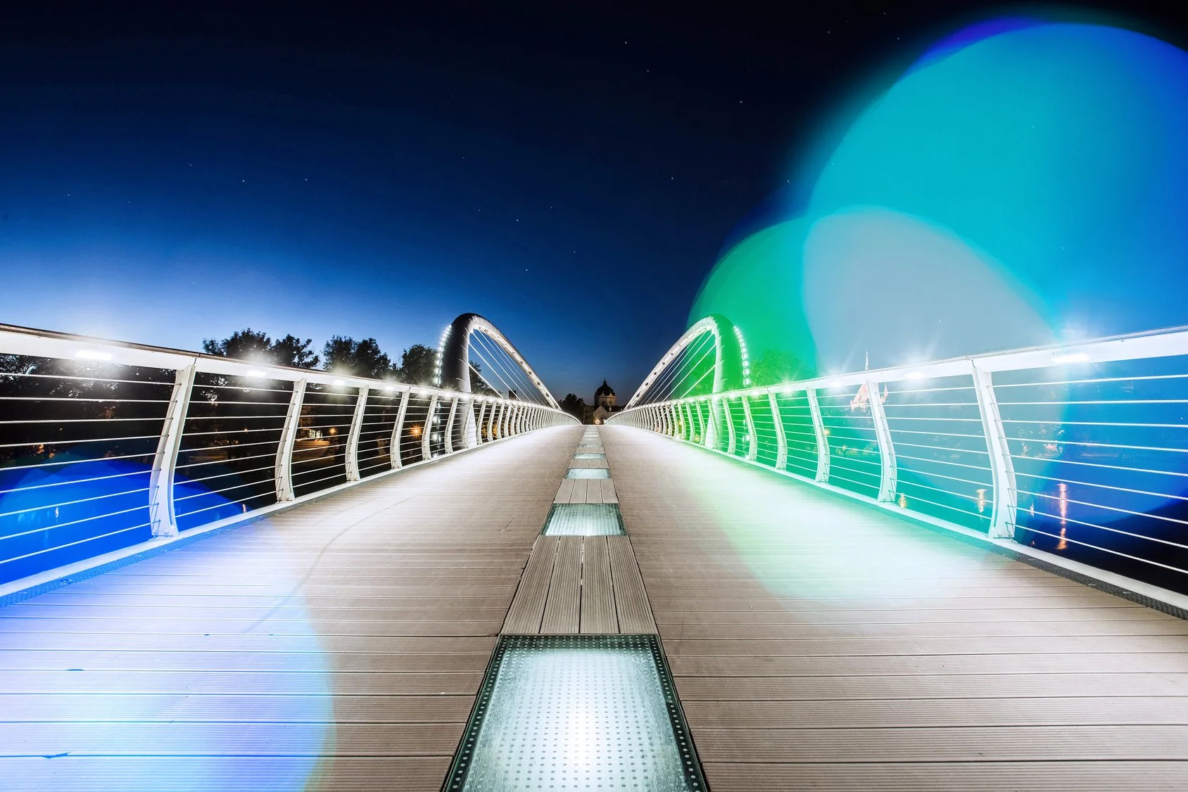 Photo of a modern illuminated pedestrian bridge at night with city lights and a dark sky in the background.