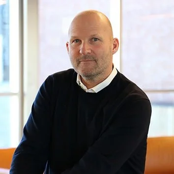 A man with a bald head and a beard sitting in an office or conference room, wearing a black sweater over a white collared shirt, near a window with a blurred cityscape background. Njål Berge.