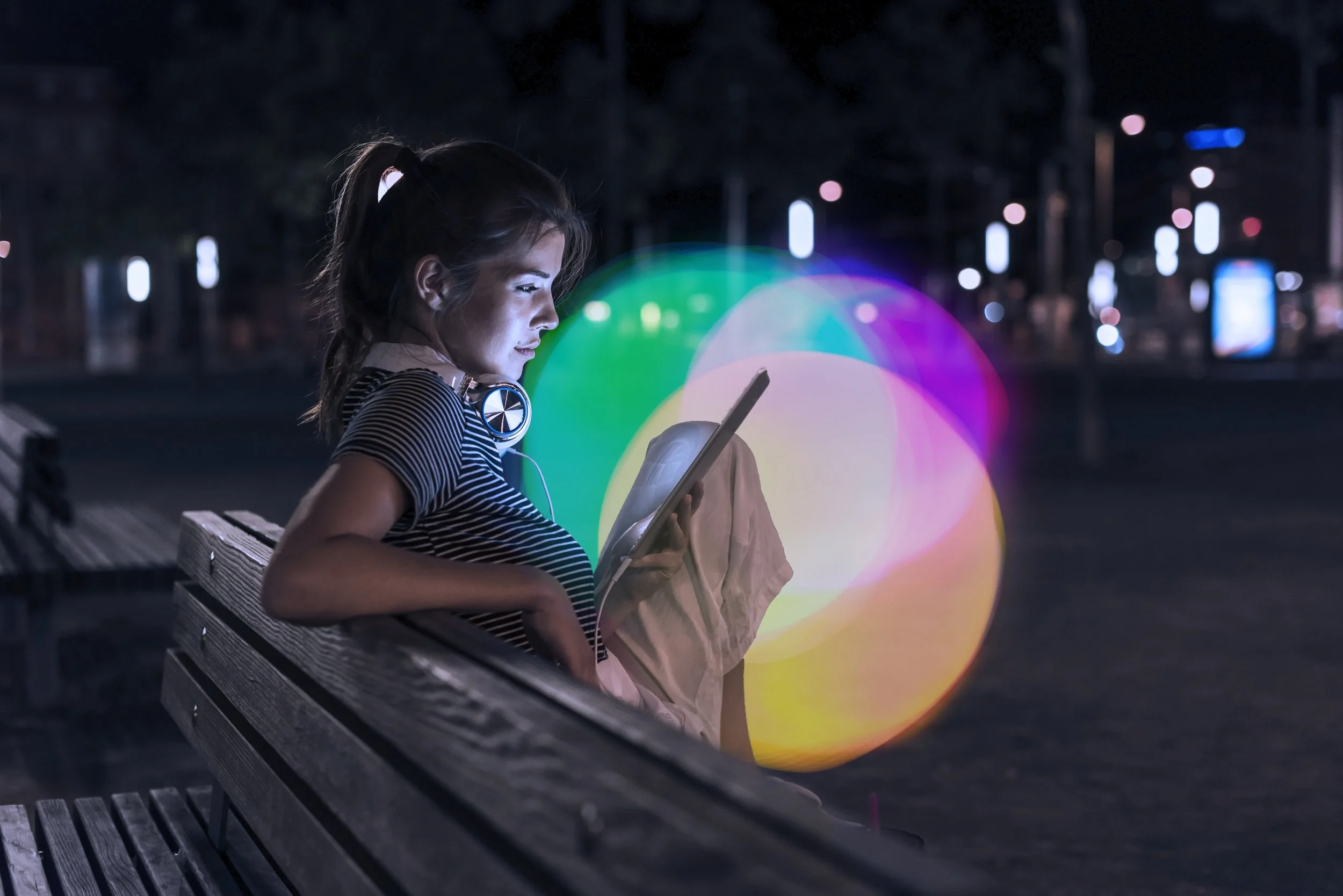 A young woman with headphones around her neck sitting on a park bench at night, looking at her tablet with colorful bokeh lights in the background.