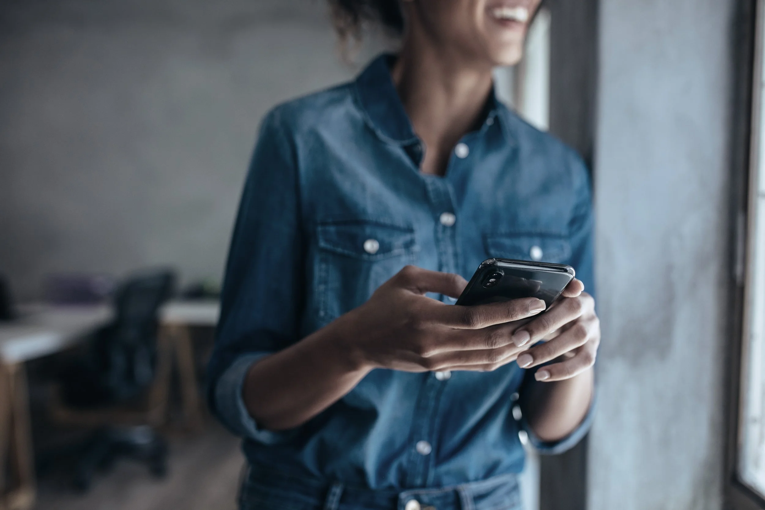 A woman standing indoors near a window, holding a smartphone in her hands, wearing a denim shirt.