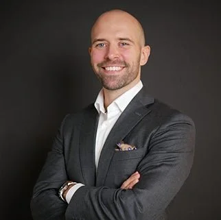 A smiling man in a dark gray suit with a white shirt, standing against a gray background. Håkon Wik.