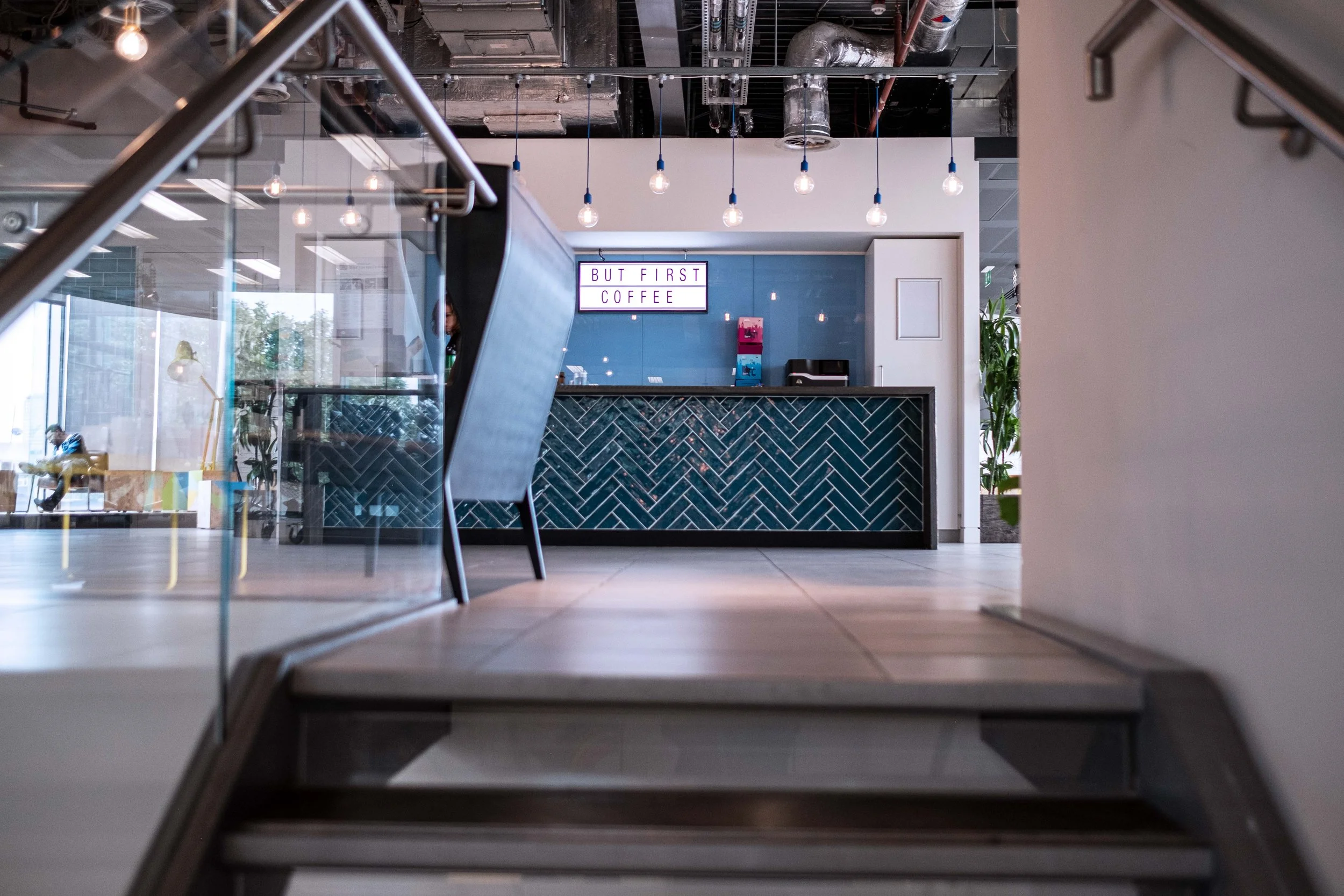 Inside a modern coffee shop with a black counter featuring a herringbone tile pattern, a sign that reads 'But First Coffee' on the wall, hanging light bulbs, and a person visible behind the counter.