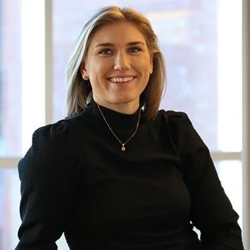 A woman with shoulder-length blonde hair smiling, wearing a black top and a necklace, standing in front of a window. Caroline Olsen.