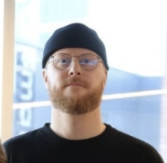 A man with glasses, a beard, wearing a black beanie and black shirt, standing indoors near a window. Christopher Axelsen.