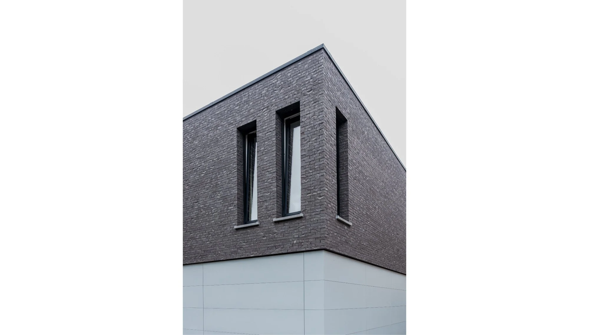 Moderne tijdloze residentiële nieuwbouwwoning met donkerbakstenen gevel en smalle raampartijen, boven een witte gevel ontworpen door Gert Anthonissen Architect..