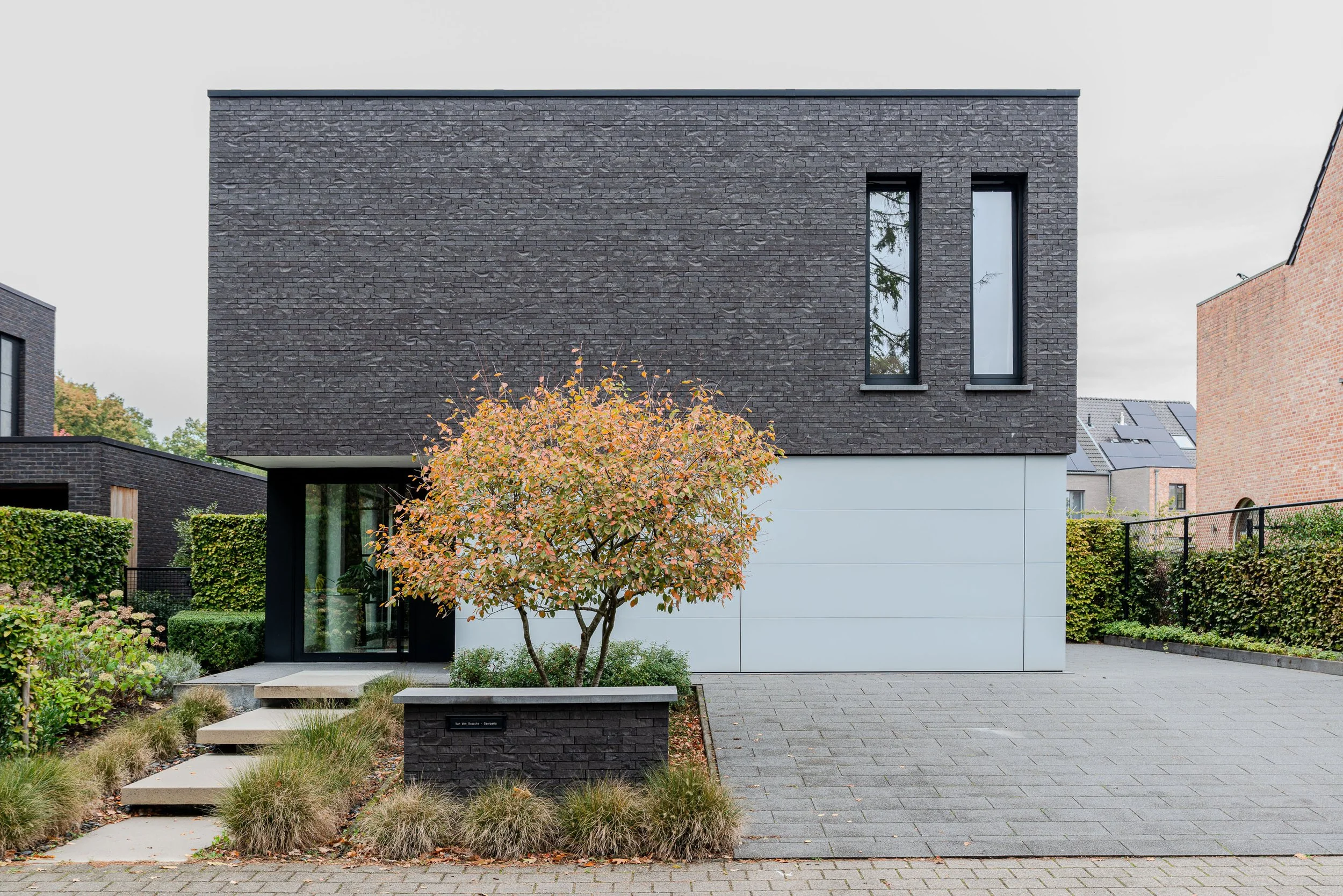 Moderne woning met zwarte en witte gevels en grote ramen, voor- en zijtuin met boom en grindpad.