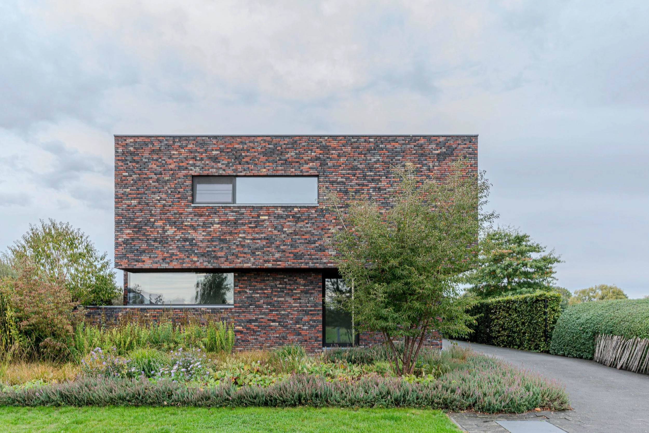 Moderne woning met een gevel van gemêleerd baksteen en grote ramen, omgeven door een tuin met diverse struiken en gras, onder een bewolkte hemel.
