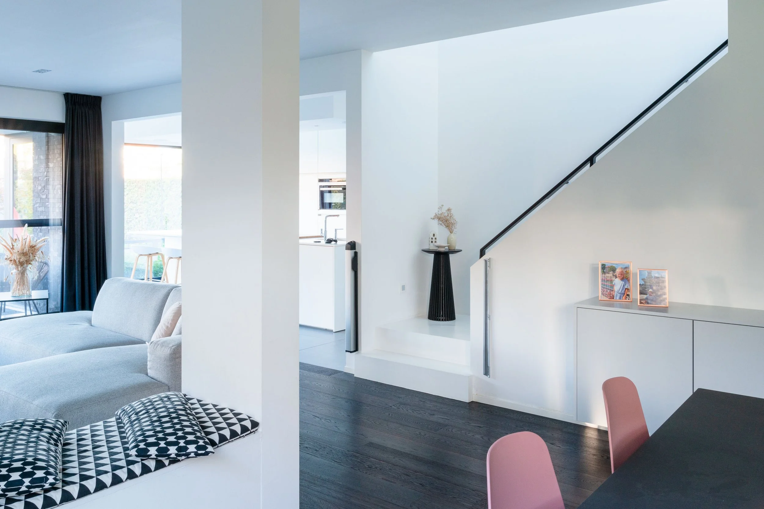 Interieur van een modern, licht en minimalistisch woonhuis met witgekleurde muren en donkere houten vloeren. Een witte bank in de woonkamer, een eettafel met roze stoelen, en decoratieve elementen zoals foto’s en vazen. Gert Anthonissen Architect.