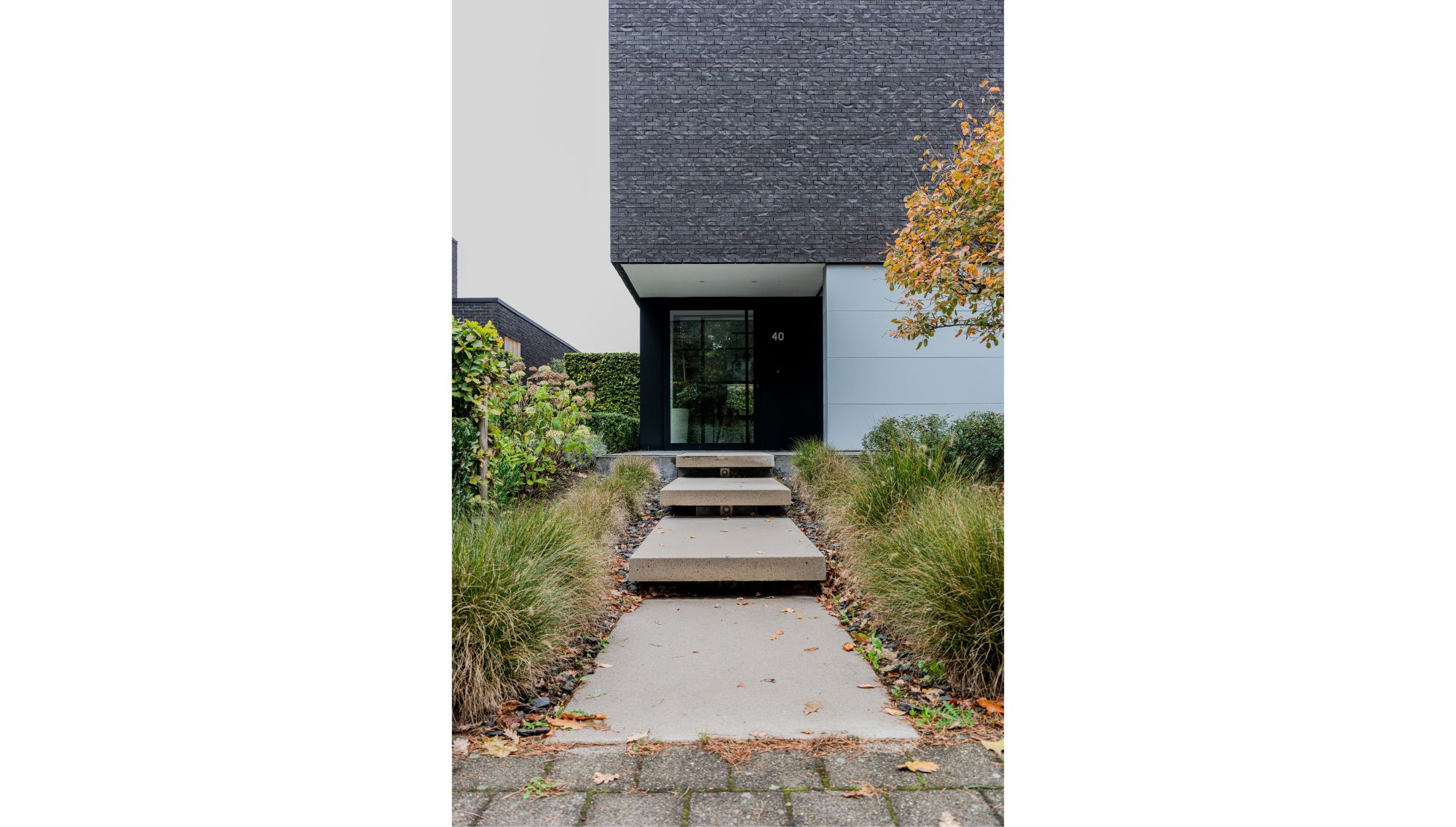 Een modern huis met een ingang en vier betonnen traptreden, omgeven door groene planten en een pijnboom in aangelegde tuin, ontworpen door Gert Anthonissen Architect. Brecht