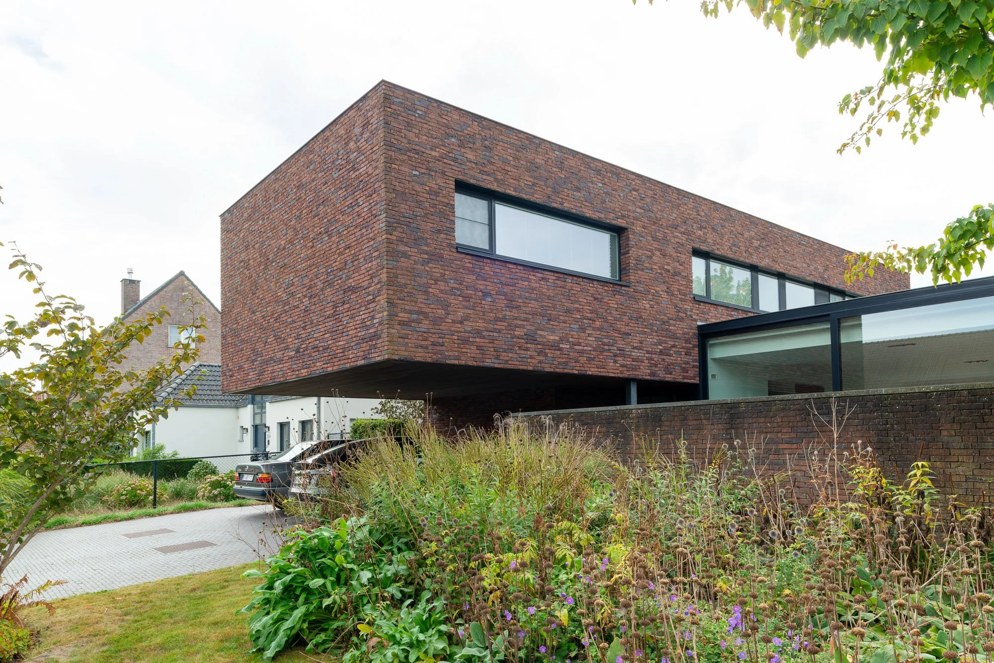 Modern huis met een rood bakstenen bovenverdieping die uitsteekt boven de onderverdieping, grote raampartijen en een tuin met bloemen en planten. Ontworpen door Gert Anthonissen Architect. Regio Loenhout