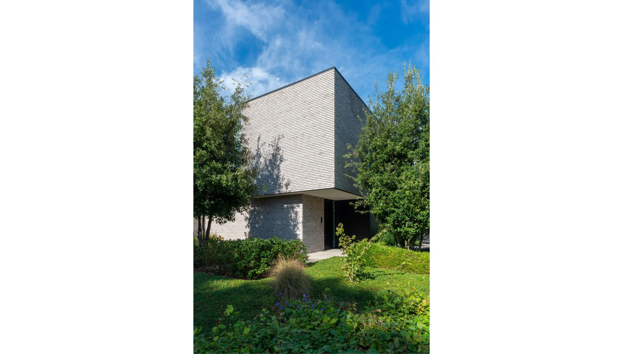 Moderne witte woning met strakke lijnen, omringd door groene struiken en bomen onder een blauwe hemel. Ontworpen door architectenbureau Gert Anthonissen Architectuur.