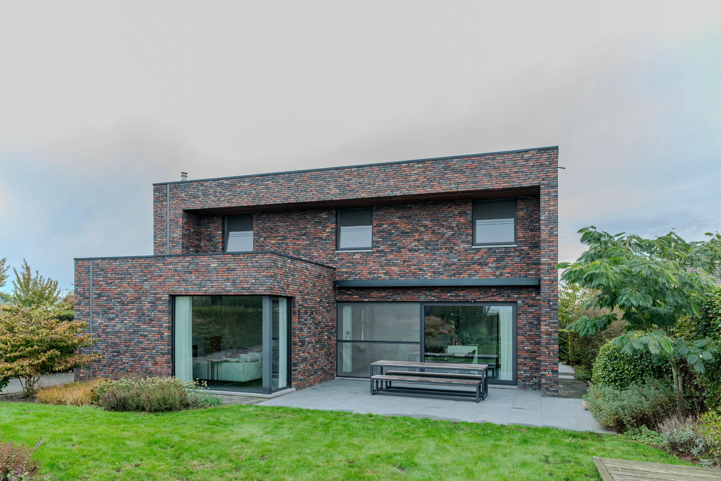 Moderne, multi-verdiepings huis met gevel van donkere bakstenen, grote glasramen, tuin met gras en bomen, patio met tafel en banken.