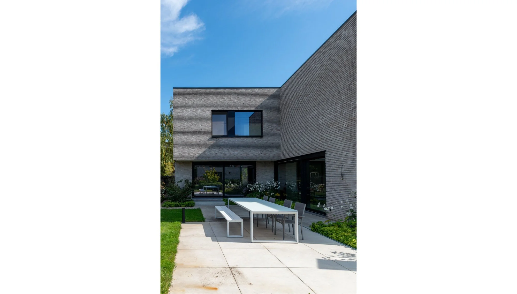 Moderne, twee verdiepingen tellende woning met een patio en buitenmeubels, omgeven door groene tuinen en een heldere blauwe lucht. Getekend door architectenbureau Gert Anthonissen Architectuur.