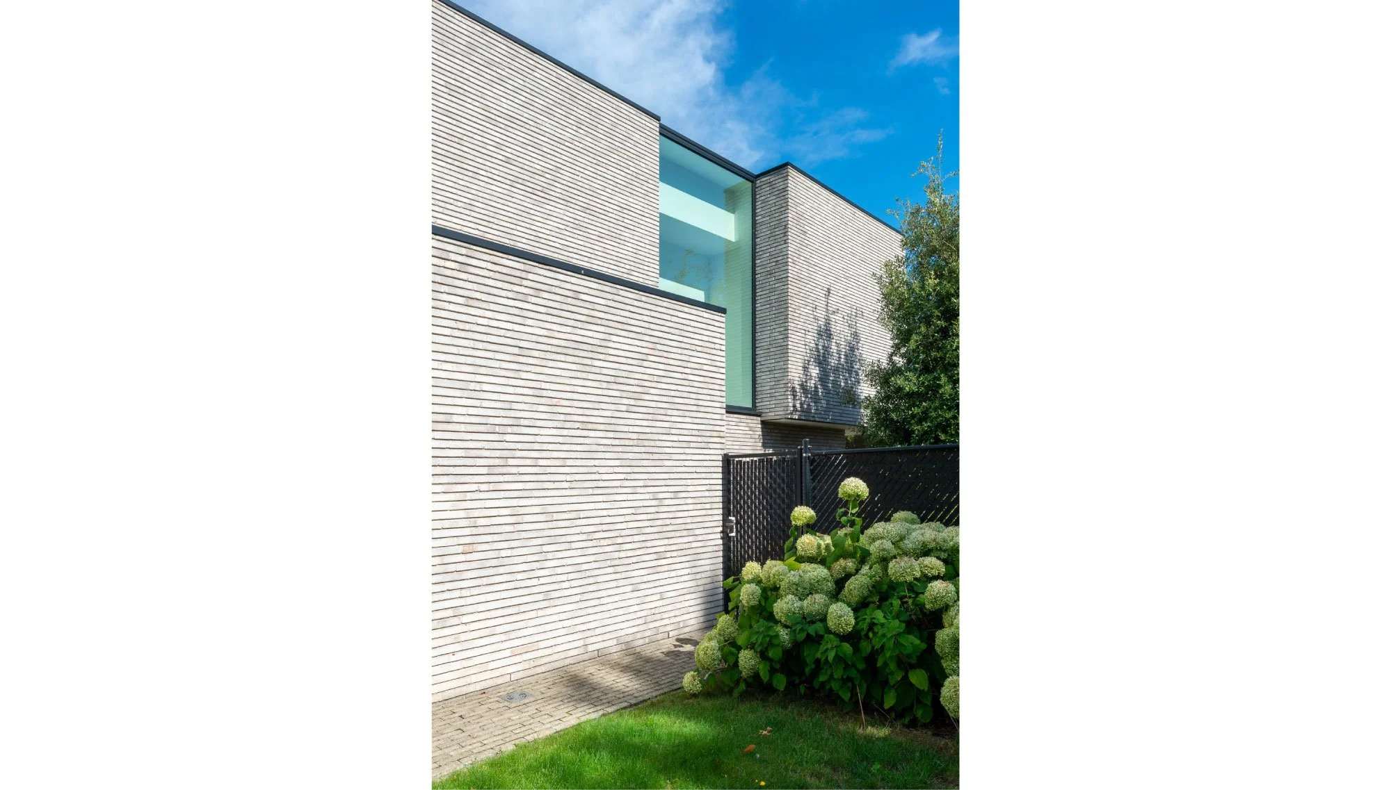 Modern nieuwbouwwoning met witte bakstenen muren, grote ramen en een tuin met groen gras, bloemen en een zwarte hek te Westmalle. Ontworpen door Gert Anthonissen Architect..
