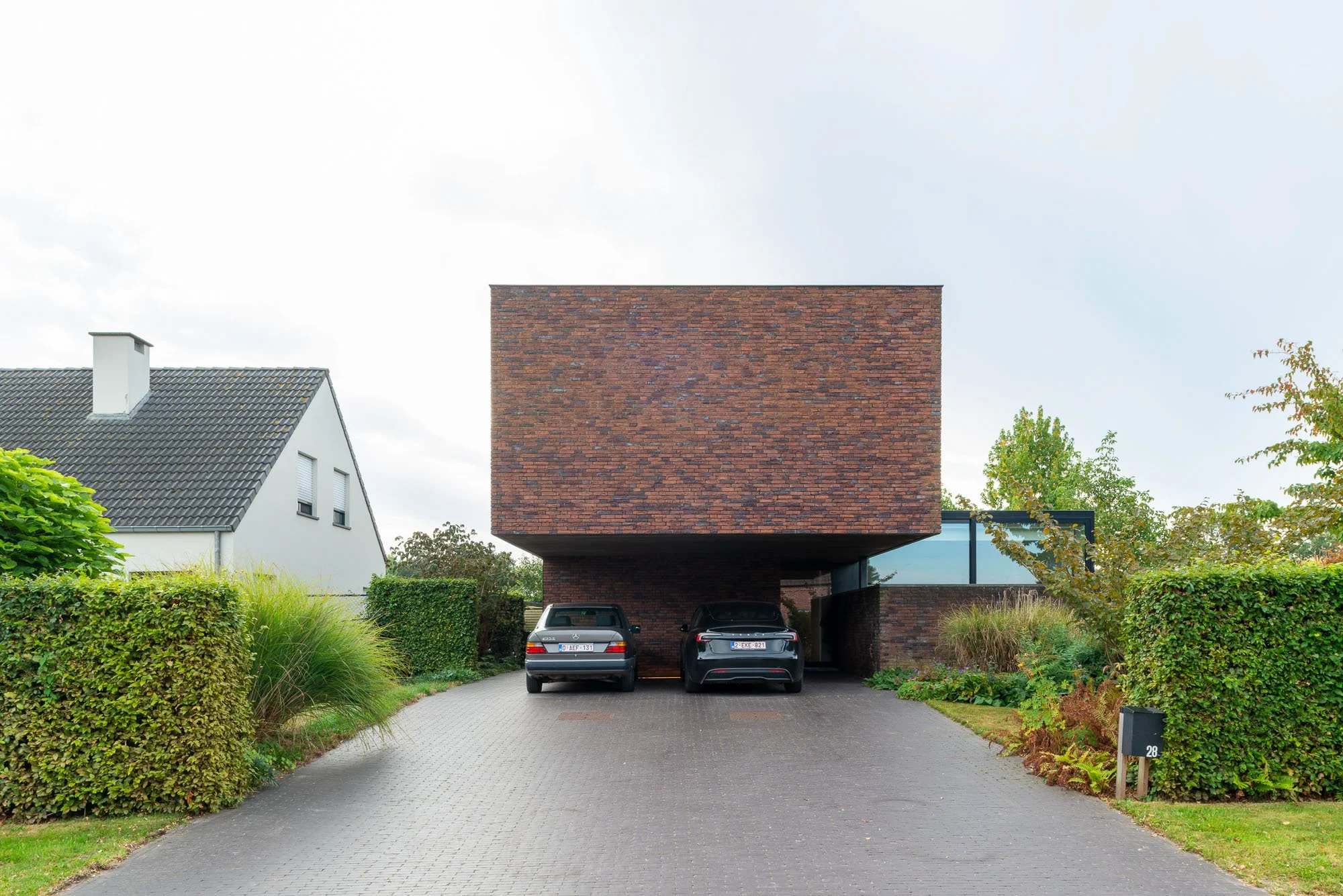 Moderne woning met een carport en twee geparkeerde auto's, omgeven door groene heggen en tuin.