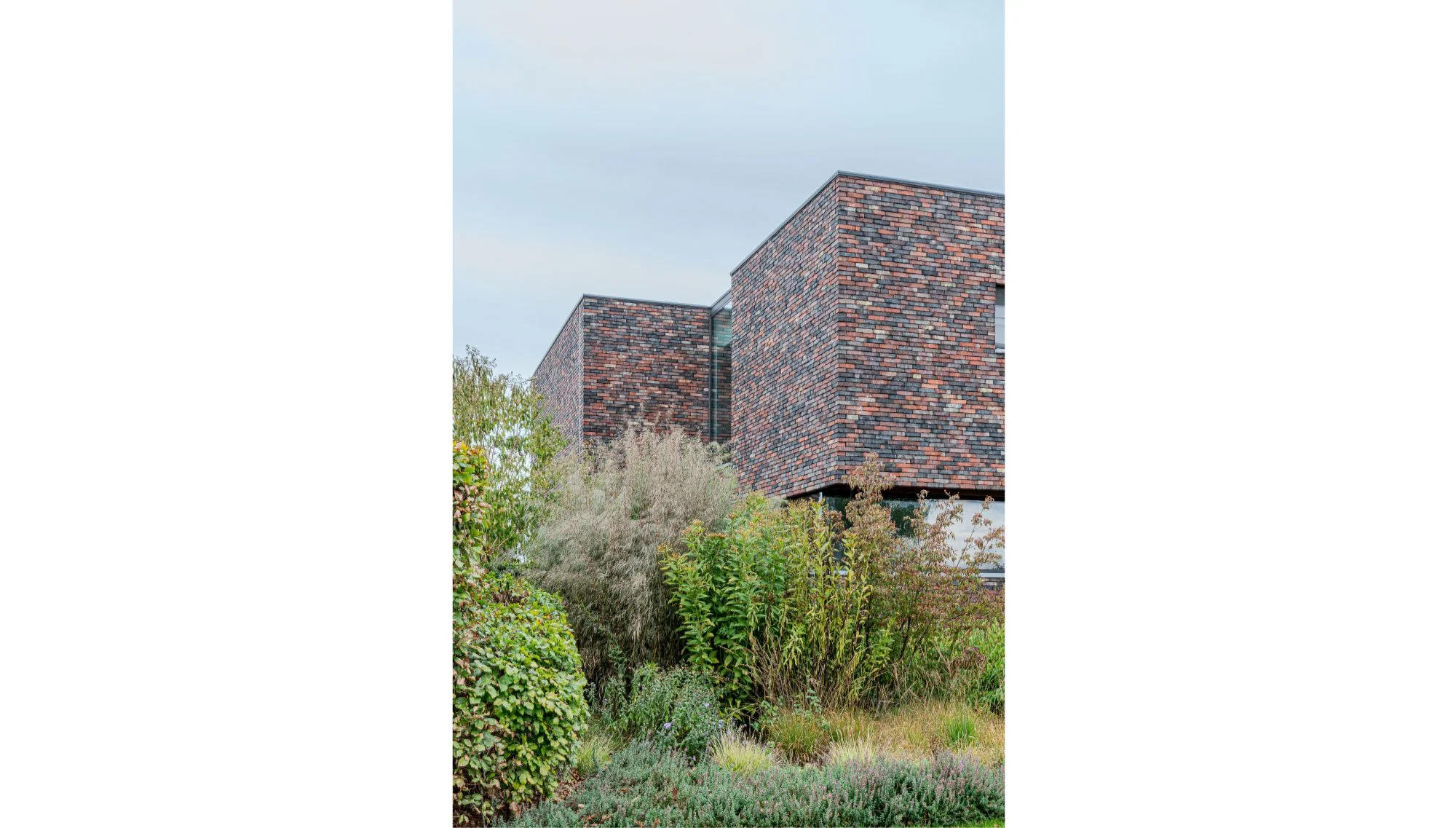 Moderne gebouw met brickwork gevel, omgeven door diverse planten en struiken, onder een bewolkte hemel. Plan ontworpen door architectenbureau Gert Anthonissen Architect regio noord Antwerpen.