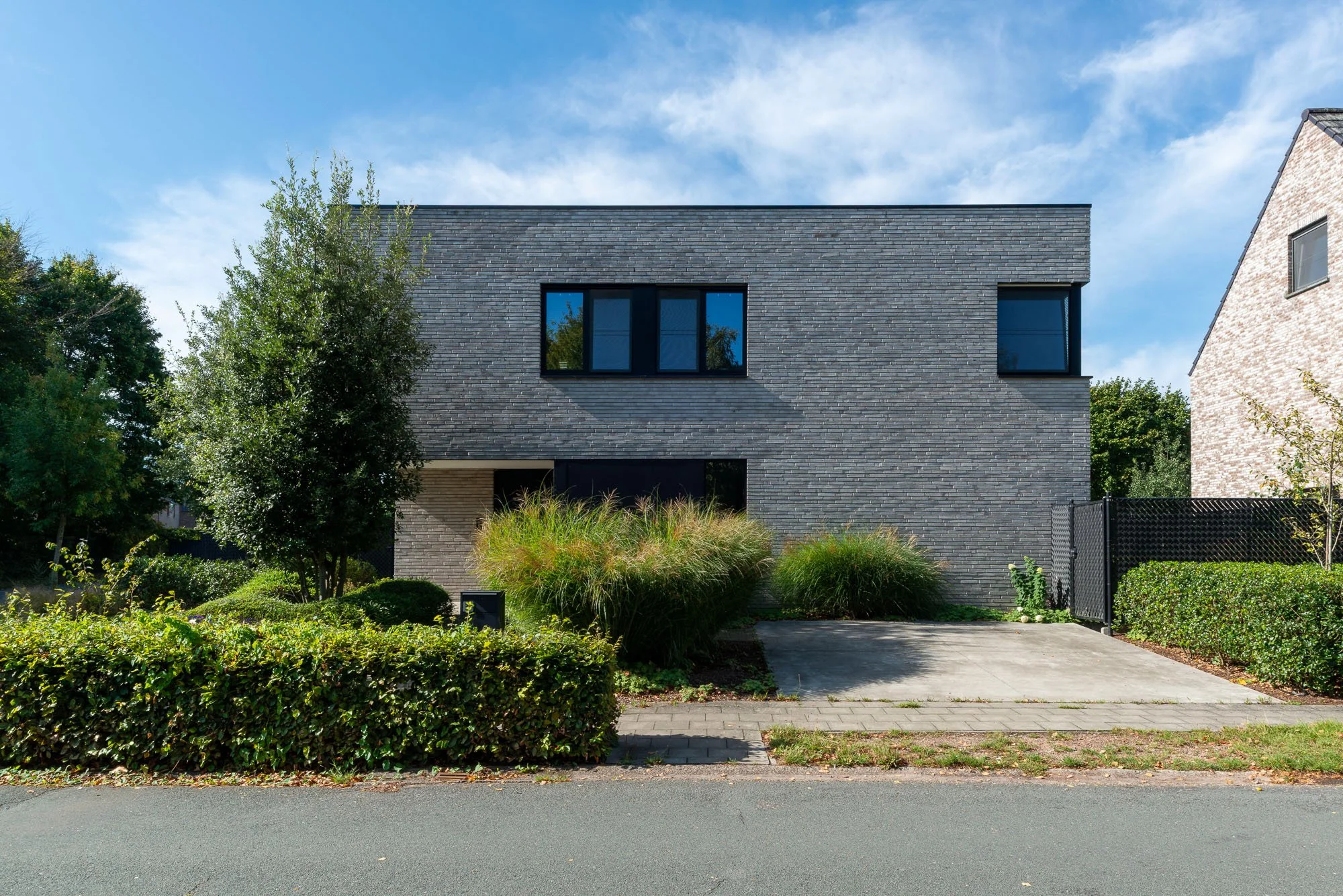 Moderne donkergrijze huis met grote ramen, omringd door groene struiken en bomen, onder een blauwe lucht met wolken.