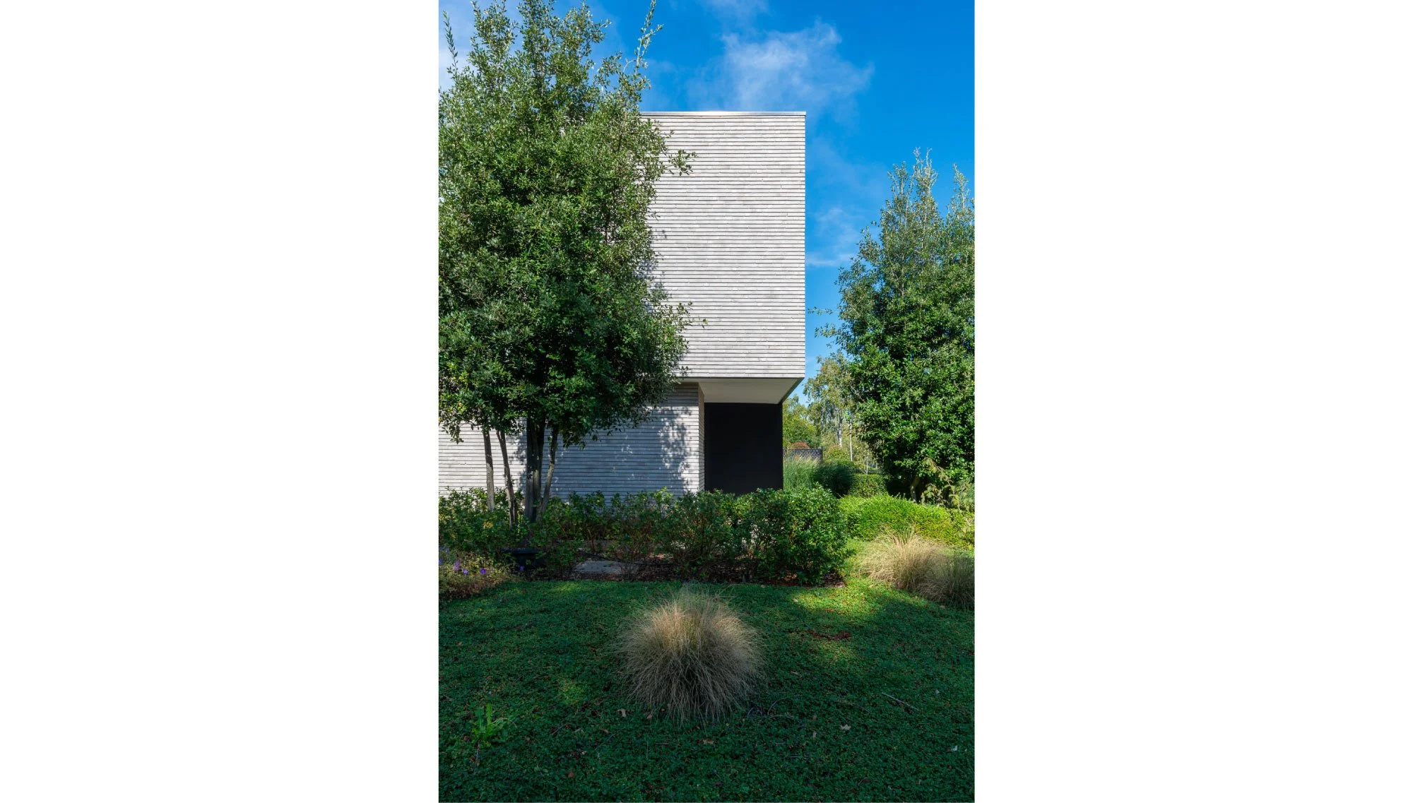 Moderne woning met witte gevel, omgeven door groene bomen en struiken, onder een blauwe hemel.