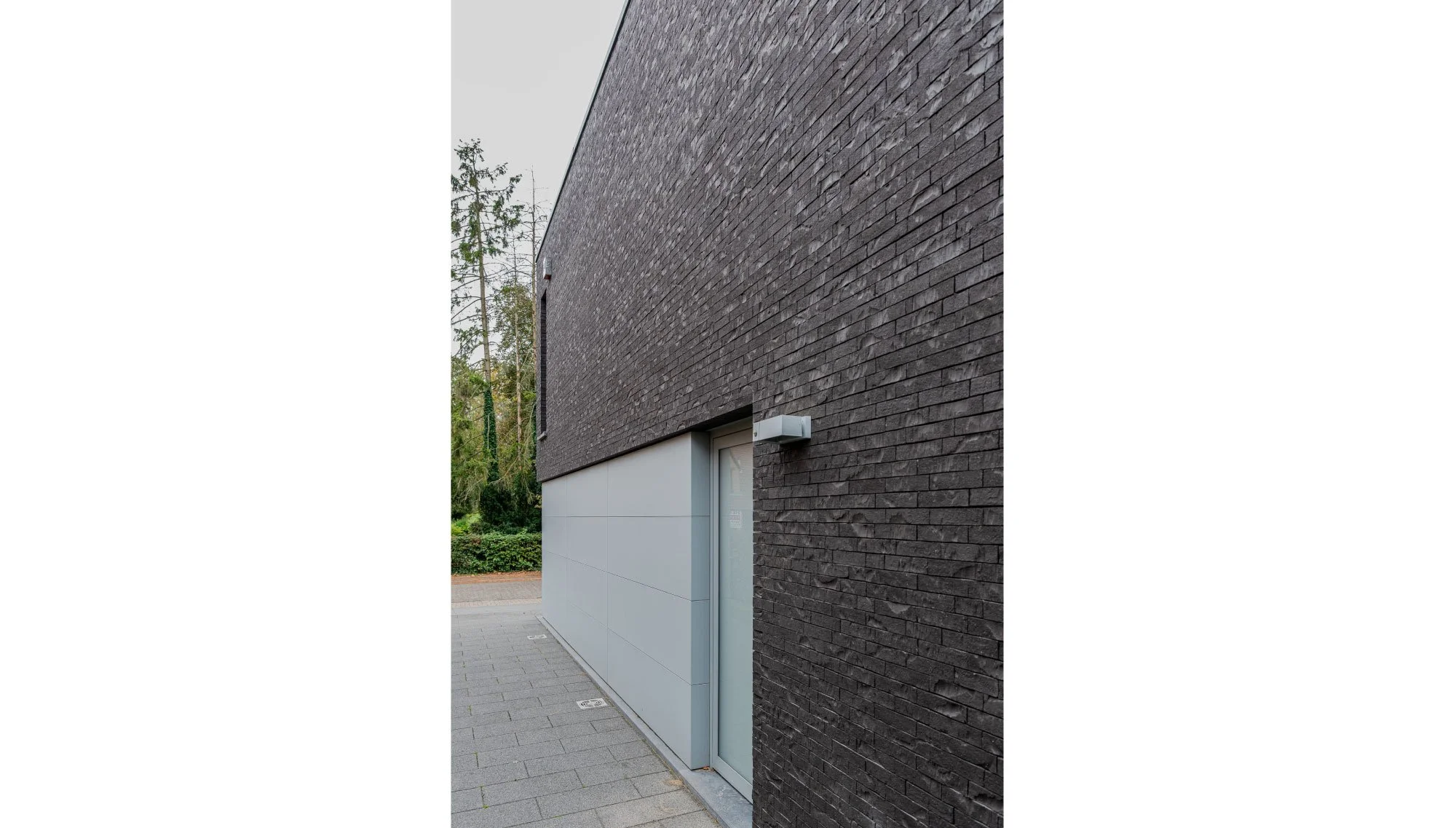 Close-up van een modern gebouw met een gevel van zwarte bakstenen en een schuifraam met gordijn erachter. Er is een lichte muur met geïntegreerde deur en buitenverlichting. Architectuur door Gert Anthonissen Architect.